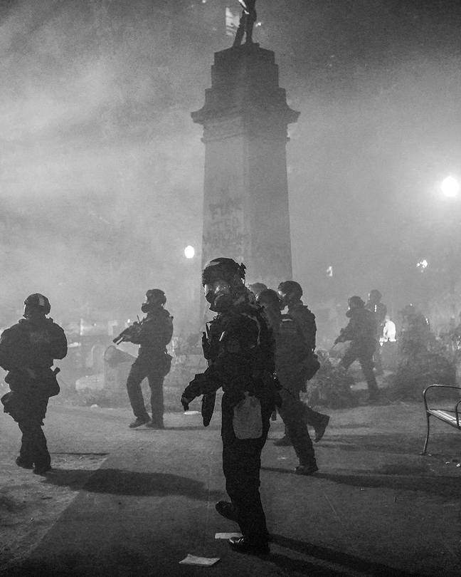 Black-and-white photo of armed troopers in full body armor and gas masks in foggy city park near statue on tall pedestal