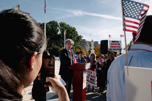 Mr. Blum speaking into a microphone at a podium outside, surrounded by supporters holding signs.