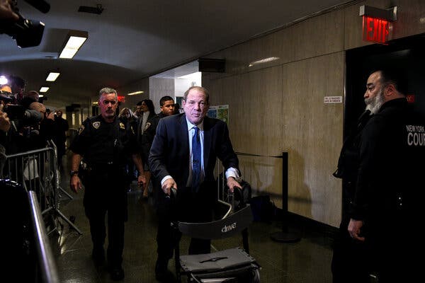 Harvey Weinstein walking through a courtroom hallway with the use of a walker, surrounded by law enforcement officers.