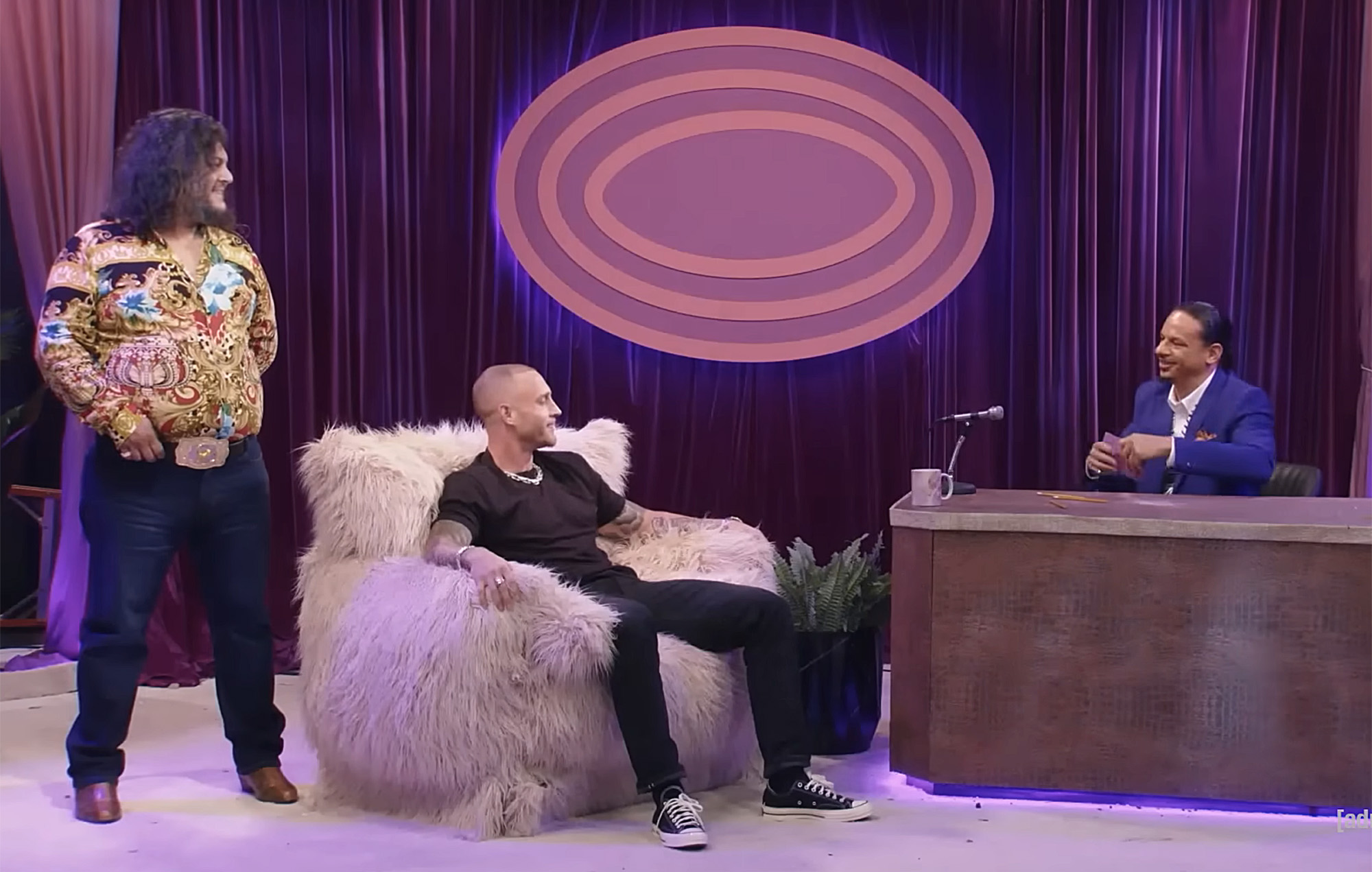 Chet Hanks sits in a fluffy armchair on the set of The Eric Andre Show, with host Eric Andre seated at a desk on the right and co-host Hannibal Buress standing on the left.