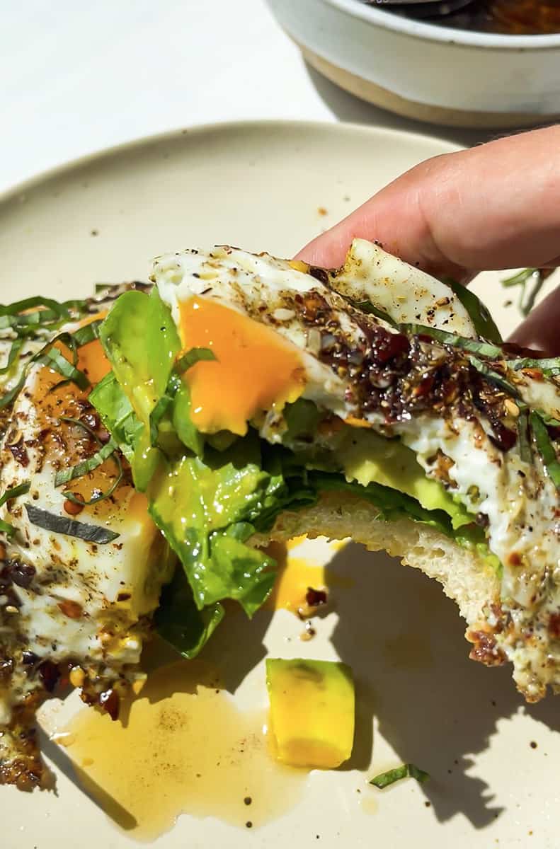 A hand holding a half piece of fried egg with spinach toast over a white bowl.