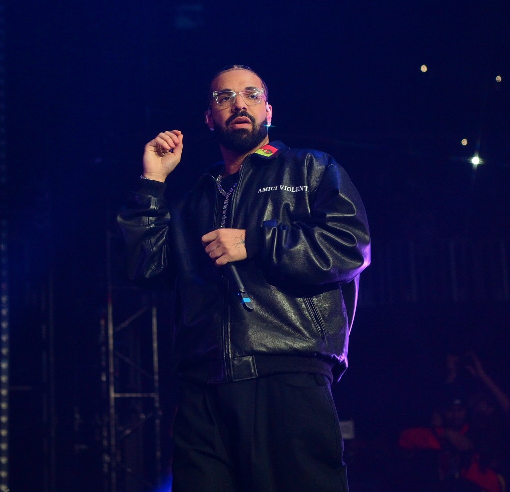 Drake stands on a stage in a leather jacket and clear glasses with a blank expression on his face