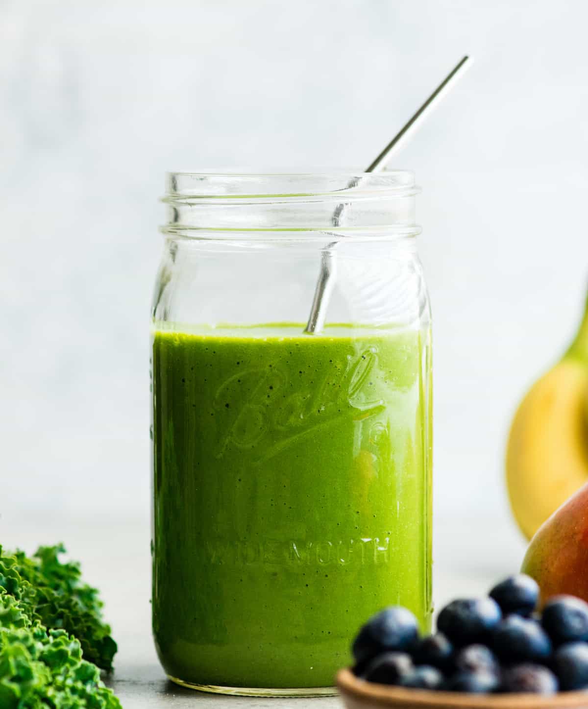 front view of a green smoothie in a large glass mason jar with a metal straw, surrounded with kale, blueberries, bananas, mangoes and green grapes