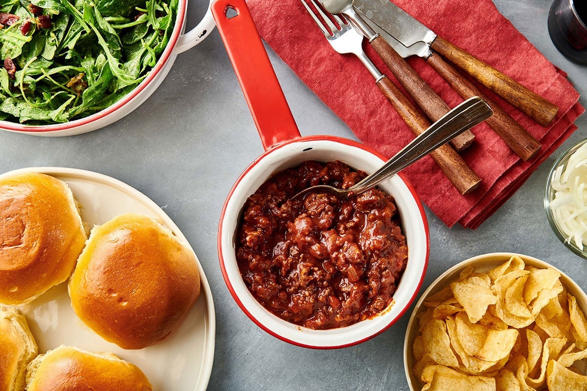 Sloppy Joe mix a pan on table with buns, chips, and salad.
