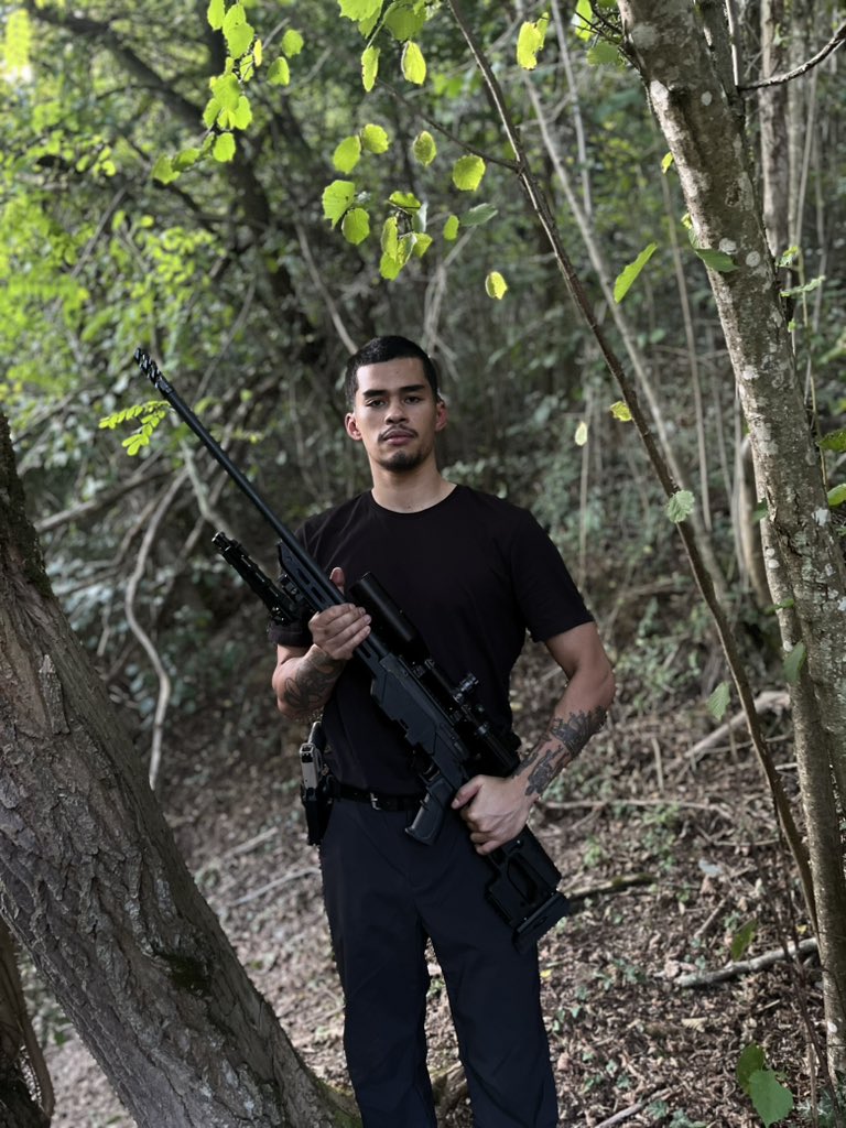 Sneako, aka Nico Kenn De Balinthazy, holding a rifle in a wooded area.