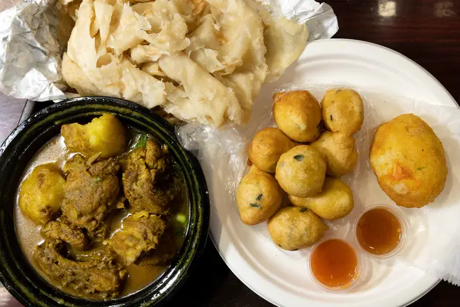 Plates of chicken curry with roti, pholourie, and egg ball are on a table at Shanta’s Bakery and Restaurant