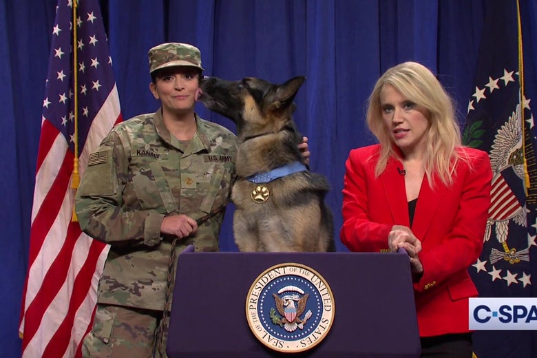 Kate McKinnon dressed as Kellyanne Conway, Cecily Strong in an Army uniform, and a German Shepherd stand behind the White House briefing room podium in a still from SNL.