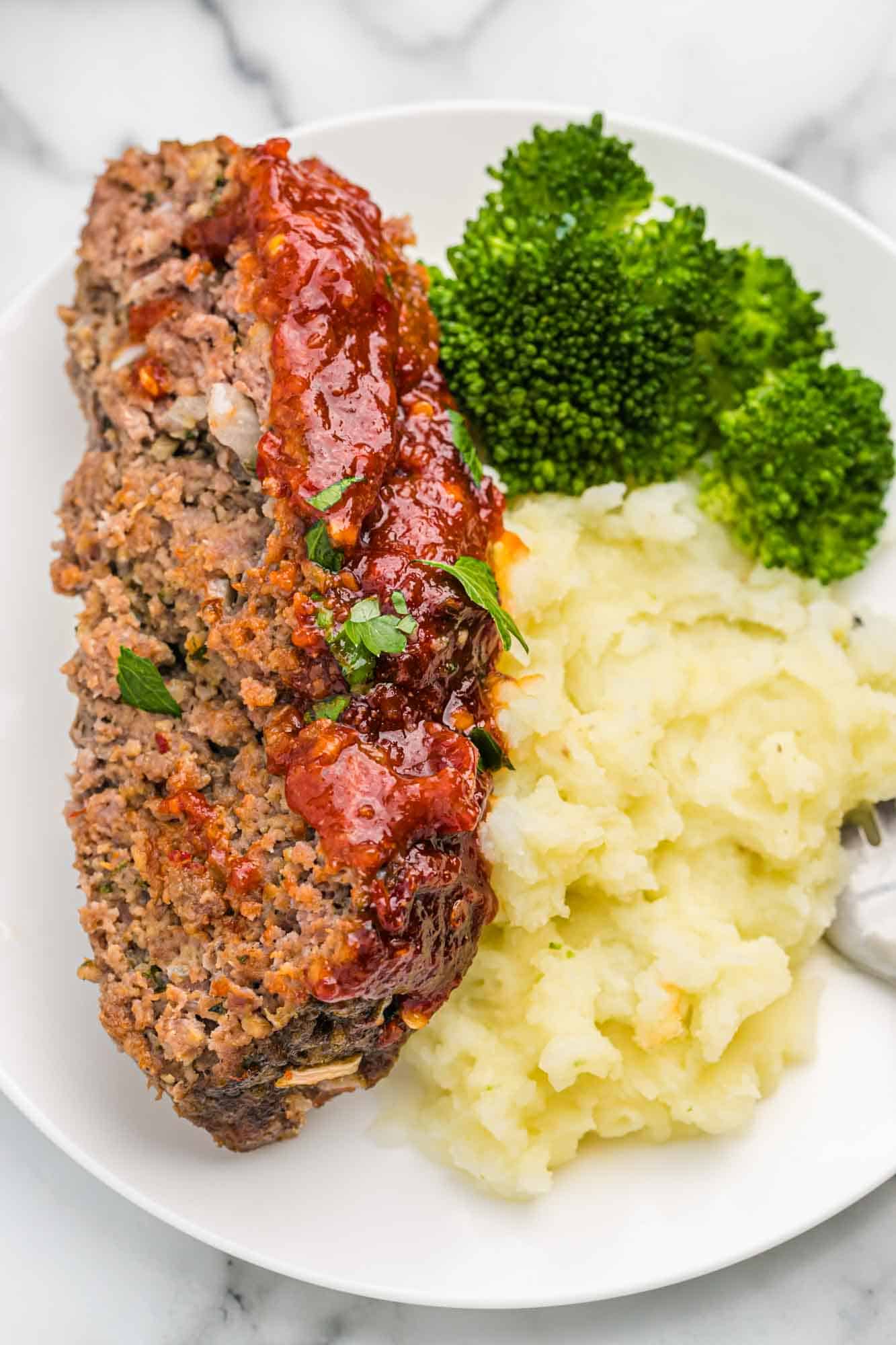 Slice of meatloaf served on a white plate with mashed potatoes and steamed broccoli